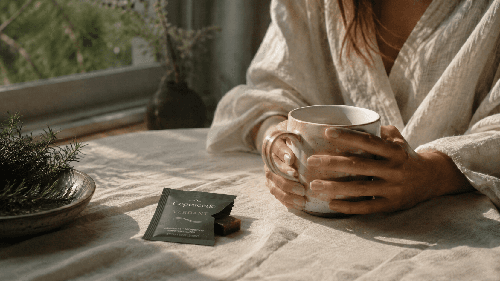 A woman at a sunlit table holding a ceramic mug, with a Copeacetic Verdant packet and rosemary sprig in the foreground